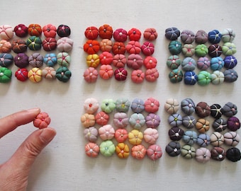 Può includere: Una collezione di bottoni colorati rivestiti in tessuto in varie tonalità di rosso, arancione, rosa, verde, blu e marrone. Un bottone è tenuto tra le dita. I bottoni sono disposti in file ordinate su una superficie bianca.