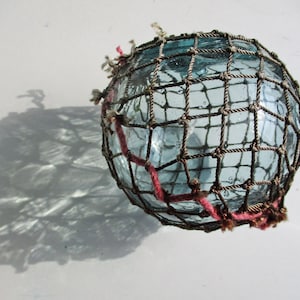 May include: A round, clear blue glass float, encased in a brown woven net. A red, knotted cord wraps around the float. The float casts a shadow on a white surface. Nautical decor.