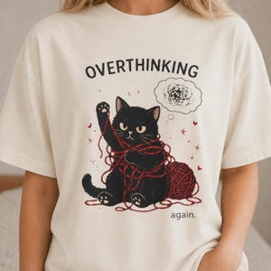 May include: A cream-coloured t-shirt with a black cat tangled in red yarn. The text reads "OVERTHINKING" above the cat and "again." below. A thought bubble with a scribbled design is above the cat's head.