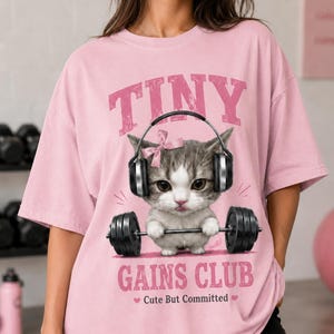 Puede incluir: Camiseta rosa claro con el texto "TINY GAINS CLUB" y un gráfico de un gatito con auriculares levantando pesas. El texto "Cute But Committed" está debajo del gráfico.
