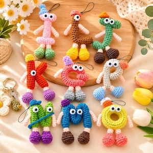 May include: A collection of nine colorful, crocheted letter-shaped keychains, each featuring googly eyes, small hats, and dangling arms and legs. The letters include pink, brown, green, red, coral, beige, lime green, blue, and yellow yarn designs.