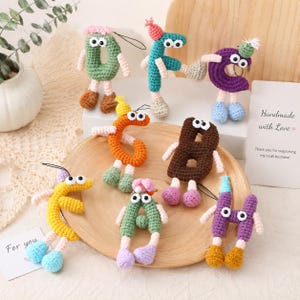 Puede incluir: Una colección de siete adornos de letras de ganchillo con ojos móviles, brazos pequeños y piernas con zapatos. Cada letra está hecha en un color diferente, incluyendo verde, turquesa, morado, naranja, marrón, amarillo y lavanda, con diminutos gorros.