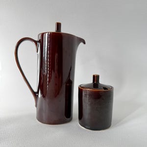 Può includere: Una caffettiera in ceramica marrone scuro con manico e beccuccio, accanto a una zuccheriera abbinata con coperchio. Il set ha una finitura lucida e un design classico, adatto per servire caffè o tè.