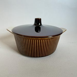 May include: A brown ceramic casserole dish with a lid. The dish has vertical ribbed detailing on the sides and a dark brown, glossy lid with a small handle. The dish has small white handles on each side.