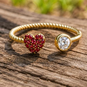 Può includere: Un bracciale aperto color oro. Un'estremità presenta una forma a cuore incastonata con piccole pietre preziose rosse. L'altra estremità ha un'unica, grande gemma trasparente. Il bracciale ha un design a corda intrecciata.