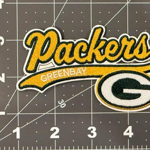 May include: An embroidered fabric patch featuring the word Packers in yellow script with a dark green outline, above the text GREENBAY and a large dark green letter G with a white outline, placed on a mat with a 5-inch ruler scale.