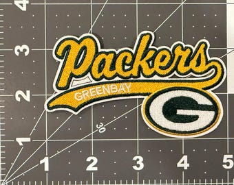 May include: An embroidered fabric patch featuring the word Packers in yellow script with a dark green outline, above the text GREENBAY and a large dark green letter G with a white outline, placed on a mat with a 5-inch ruler scale.