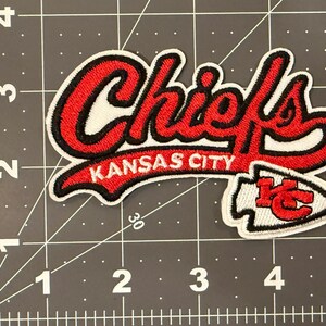 May include: An embroidered fabric patch featuring the red and white text Chiefs and Kansas City, with a red and white arrowhead logo containing the letters KC. The patch is placed on a cutting mat with inch markings, showing it is 4 inches wide.