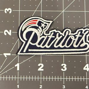 May include: An embroidered fabric patch featuring the New England Patriots logo and the word Patriots in white script. The patch is placed on a cutting mat with a grid, showing it measures approximately 4 inches in width and 2 inches in height.