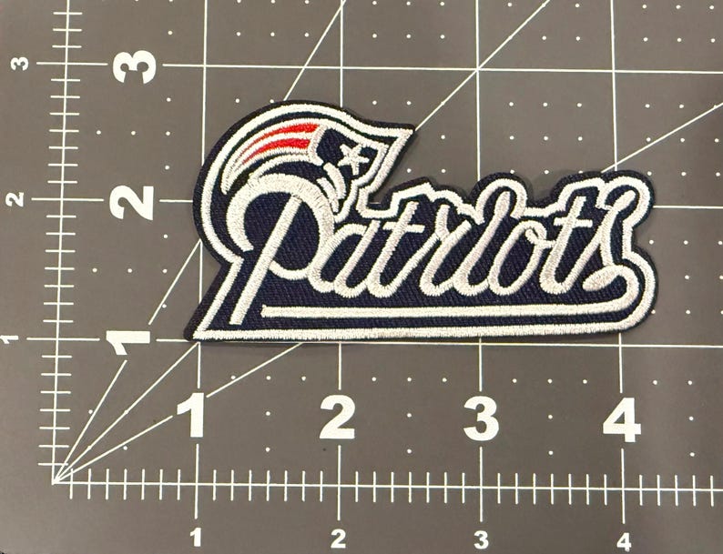 May include: An embroidered fabric patch featuring the New England Patriots logo and the word Patriots in white script. The patch is placed on a cutting mat with a grid, showing it measures approximately 4 inches in width and 2 inches in height.