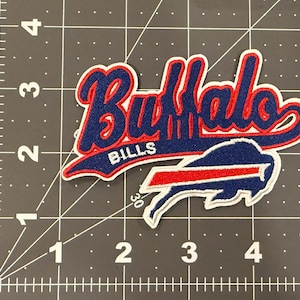 May include: An embroidered fabric patch featuring the text Buffalo Bills in blue script with a red outline and white border, positioned above a blue and red buffalo logo. The patch is placed on a cutting mat with a visible 5-inch ruler scale.