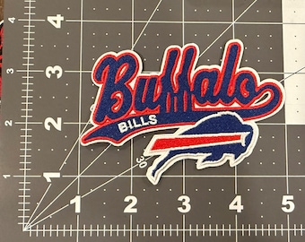 May include: An embroidered fabric patch featuring the text Buffalo Bills in blue script with a red outline and white border, positioned above a blue and red buffalo logo. The patch is placed on a cutting mat with a visible 5-inch ruler scale.