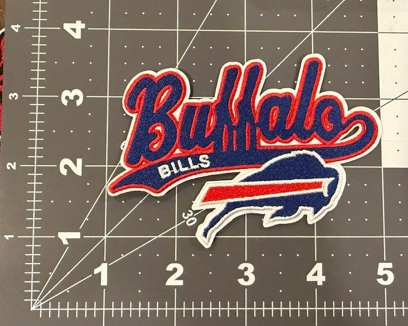 Może przedstawiać: Haftowana naszywka z tkaniny z napisem Buffalo Bills niebieską czcionką z czerwoną obw&oacute;dką i białym brzegiem, umieszczona nad logo żubra w kolorze niebieskim i czerwonym. Naszywka leży na macie do cięcia z widoczną podziałką 12,7 cm.