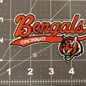 Może przedstawiać: Haftowana naszywka z tkaniny przedstawiająca słowo Bengals pomarańczową kursywą z czarnym obrysem, słowo Cincinnati w kolorze białym oraz grafikę głowy tygrysa. Naszywka umieszczona jest na macie do cięcia z oznaczeniami w calach.