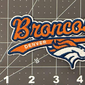 May include: An embroidered fabric patch featuring the word Broncos in orange script with a dark blue outline, positioned above the word Denver and a stylized white and orange horse head logo, measuring approximately 4.5 inches in width.