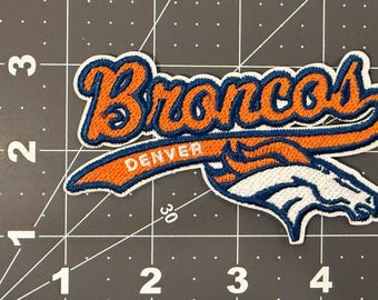 May include: An embroidered fabric patch featuring the word Broncos in orange script with a dark blue outline, positioned above the word Denver and a stylized white and orange horse head logo, measuring approximately 4.5 inches in width.