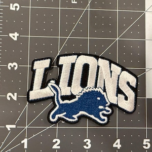 May include: A fabric patch featuring the word LIONS in white embroidered letters with a black border. Below the text is a blue embroidered lion silhouette with a white outline. The patch is placed on a cutting mat with inch markings.