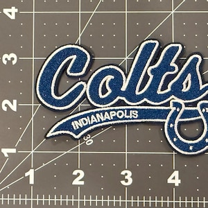 May include: A blue and white embroidered patch featuring the word Colts in a script font, with the word Indianapolis and a horseshoe icon below. The patch is positioned on a cutting mat with inch markings, showing a width of 4.5 inches.