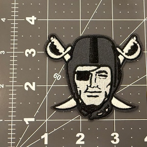 May include: An embroidered patch featuring a stylized figure wearing a football helmet and an eye patch, positioned over two crossed swords. The patch is placed on a cutting mat with visible inch markings, showing it is approximately 3 inches wide.