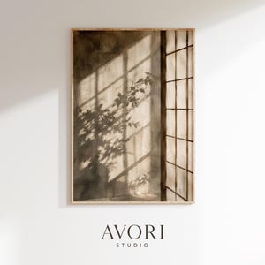 May include: A rectangular wall art print in a light wood frame featuring a sepia-toned watercolor painting of a window with a grid pattern casting long shadows of leafy plant branches onto a wall. The text AVORI STUDIO appears below the print.