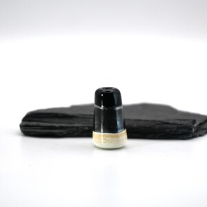 Puede incluir: Un tapón de botella de cerámica en blanco y negro con una base color crema. El tapón es cilíndrico y se encuentra frente a una piedra rectangular oscura y texturizada sobre un fondo blanco.