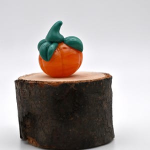 May include: A miniature orange pumpkin figurine with green leaves and stem, displayed on a small, round wooden base. The pumpkin is a vibrant orange color, and the leaves are a deep green. The wooden base has a natural, rustic appearance.