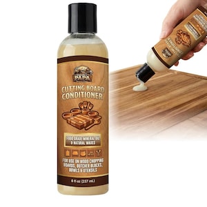 Puede incluir: Una botella de acondicionador para tablas de cortar con tapa negra y etiqueta marrón. La etiqueta dice "Project Papa Cutting Board Conditioner" y enumera ingredientes y usos. La botella está junto a una tabla de cortar de madera.