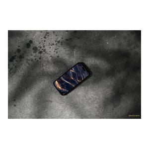Könnte beinhalten: Ein schwarzes Smartphone mit einem Bildschirm, der ein abstraktes Design in Dunkelblau und Gold zeigt. Das Telefon liegt diagonal auf einer strukturierten, dunkelgrauen Oberfläche. Das Bild hat einen Schatteneffekt.