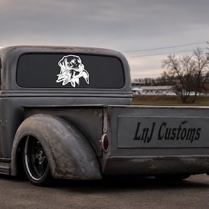 May include: A white vinyl decal featuring a stylized illustration of a hunting dog holding a duck in its mouth, applied to the rear window of a gray vintage pickup truck with the text LnJ Customs visible on the tailgate of the vehicle.