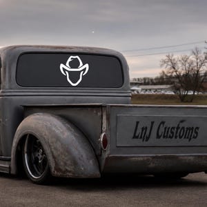 May include: A white vinyl decal featuring a stylized cowboy hat icon applied to the rear window of a gray vintage pickup truck. The truck tailgate displays the black text LnJ Customs in a cursive font, set against a weathered metal finish.