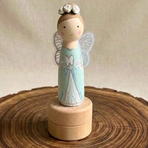 May include: A wooden fairy figurine with a pale blue dress, white wings, and a flower crown. The fairy is on a small, round wooden box. The figurine has a painted face with rosy cheeks and a gentle smile. The background is a light, neutral colour.