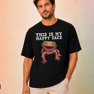 May include: A black short-sleeved t-shirt featuring a graphic of a bearded dragon with an open mouth. Above the lizard, the text reads THIS IS MY HAPPY FACE in white capital letters. The shirt has a crew neckline and a relaxed fit.