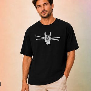 May include: A black short-sleeved t-shirt featuring a white graphic print of a hand making a rock and roll sign while holding two drumsticks. The design has a distressed, textured appearance and is centred on the chest of the garment.