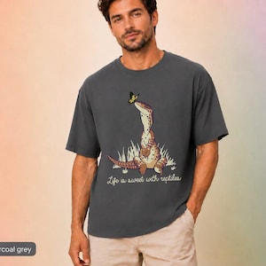 May include: A dark charcoal grey t-shirt featuring a graphic of a spotted lizard sitting in grass with a yellow butterfly on its nose. Below the lizard, the text reads, "Life is sweet with reptiles" in a cursive, light-coloured script font.