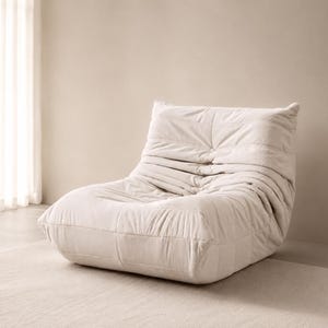 May include: A cream-coloured, modern lounge chair with a low profile. The chair features a quilted design with horizontal and vertical stitching, creating a textured appearance. The chair is made of a soft, plush material, designed for comfort and relaxation.