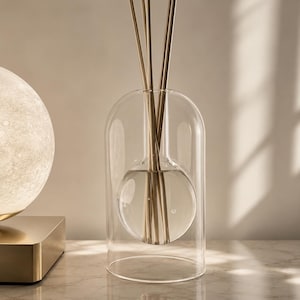 May include: A clear glass reed diffuser featuring a double-walled design with a spherical inner chamber and a cylindrical outer shell. Several thin, light-coloured wooden reeds are inserted into the centre, extending upwards from the liquid.