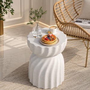 Peut inclure: Une table d'appoint blanche et texturée avec un design superposé unique. La table est garnie d'une assiette de crêpes, de fruits frais et d'une tasse de café. Un vase transparent avec de la verdure et une chaise en osier sont à proximité.