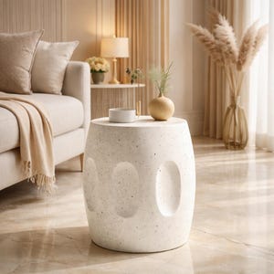 Peut inclure: Une table d'appoint en forme de tonneau, de couleur blanc cassé moucheté, avec trois découpes ovales. La table est surmontée d'un petit bol rond blanc, d'un petit vase avec de la verdure et d'un vase beige sphérique.