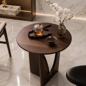 Peut inclure: Une table d'appoint ronde en bois brun foncé avec un piètement en forme de V. Une théière en verre avec une poignée en bois repose sur un plateau noir, à côté d'un petit bol. Un vase d'orchidées blanches et un vase décoratif sont également sur la table.