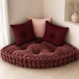 Peut inclure: Un grand coussin de sol rond et capitonné en tissu texturé bordeaux profond. Deux coussins carrés bordeaux avec des boutons centraux sont posés dessus, accompagnés d'un coussin carré rose clair lisse placé au centre de l'ensemble.