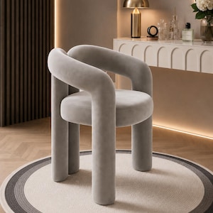May include: A contemporary, pale grey upholstered chair with curved arms and legs. The chair is placed on a circular rug with grey and white concentric rings. A gold lamp and vanity are in the background.