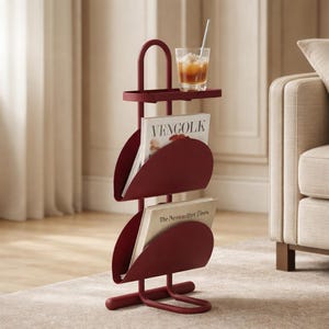 May include: A burgundy metal magazine rack with a small tray and curved magazine holders. The rack holds two magazines, one with the text "VENGOLK" and the other with "The New York Times". A glass of iced beverage sits on the tray.