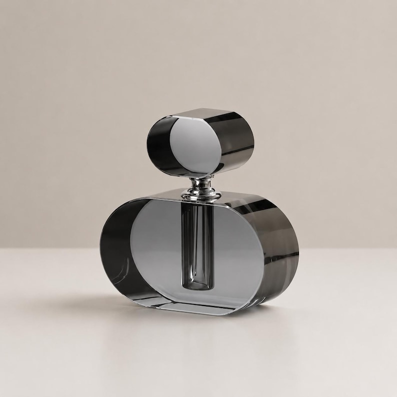 May include: A decorative glass vase featuring a wide, oval-shaped base and a smaller, matching oval-shaped top piece. The object is crafted from translucent, dark gray tinted glass with a polished, reflective finish and a central cylindrical tube.