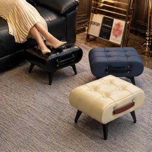 May include: Three tufted ottomans in black, navy blue, and cream. Each has a handle and sits on four black legs. The black ottoman has a person's legs resting on it. The ottomans are on a patterned rug.