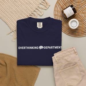 May include: A navy blue short-sleeved t-shirt with a crew neckline. The centre of the chest features white text that reads OVERTHINKING DEPARTMENT with a small white graphic of a human brain positioned between the two words.
