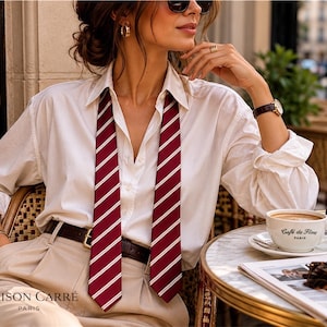 Puede incluir: Una mujer con camisa blanca, pantalones beige y corbata a rayas burdeos y blancas. Está sentada en una mesa de café con una taza de café, un periódico y gafas de sol. Un bolso burdeos está sobre la mesa.