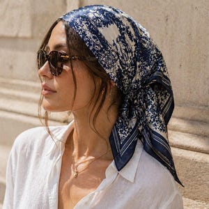 May include: A woman wearing a navy and white patterned silk headscarf. The scarf has a floral design. She is also wearing tortoiseshell sunglasses and a white button-down shirt. The scarf is tied at the back.