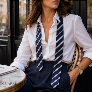 Può includere: Una camicia bianca e pantaloni blu navy sono abbinati a una cravatta a righe con strisce bianche su sfondo blu navy. È visibile un orologio da polso dorato. La scena è ambientata in un caffè, con il testo "MAISON CARRÉ PARIS" in basso.