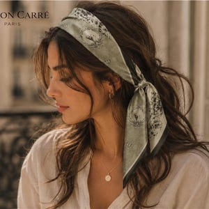 Könnte beinhalten: Eine Frau mit einem Seidenschal als Haarband. Der Schal ist olivgrün mit einem Blumen- und Wappenmuster. Der Text "MAISON CARRÉ PARIS" ist sichtbar. Sie trägt eine goldene Halskette und Creolen.