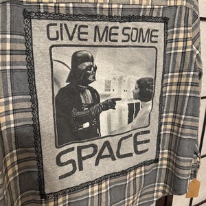 May include: Gray plaid flannel shirt with a graphic featuring Darth Vader and Princess Leia with the text "GIVE ME SOME SPACE." The graphic is framed with black lace.